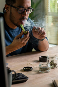 Man smoking a cannabis from smoking pipe, with cannabis equipment and accessories on the table.