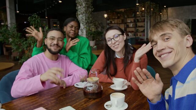 POV of company of four cheerful multiethnic friends waving and looking at camera while recording social media video in cafe