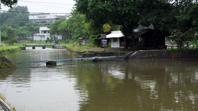 Murky brown river water flowing towards Siman Reservoir on an overcast afternoon, passing rural buildings and water pipes in Kediri, Indonesia, Southeast Asia.