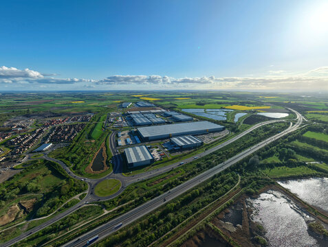 Aerial view of the iPort Avenue logistics hub with large warehouses, surrounding green fields, and a motorway under a clear blue sky in Loversall, England, United Kingdom.