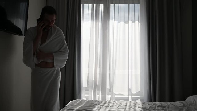 Stressed young businessman in a bathrobe pacing back and forth while talking on his mobile phone in a hotel bedroom, appearing concerned and worried before sitting on the bed
