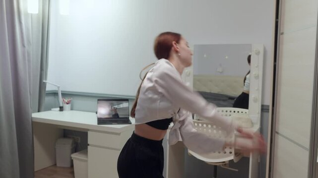 Young energetic woman with red hair dances in her bedroom. She follows an online tutorial on her laptop, learning new moves. Modern lifestyle, home workout, and remote learning concept