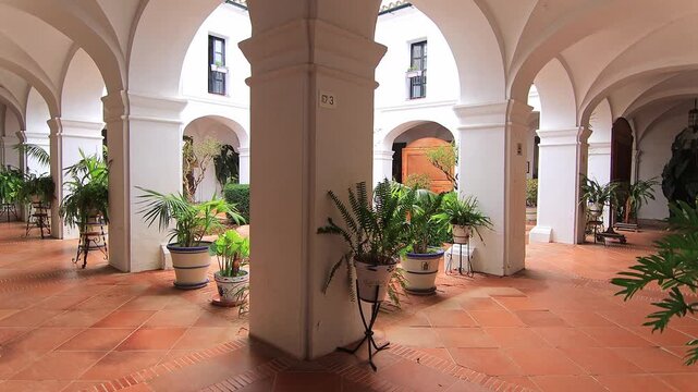Interior view of the peaceful Mudejar style cloister at Monasterio de la Rabida, featuring white arches, terracotta tiles, and lush potted plants. Palos de la Frontera, Spain.
