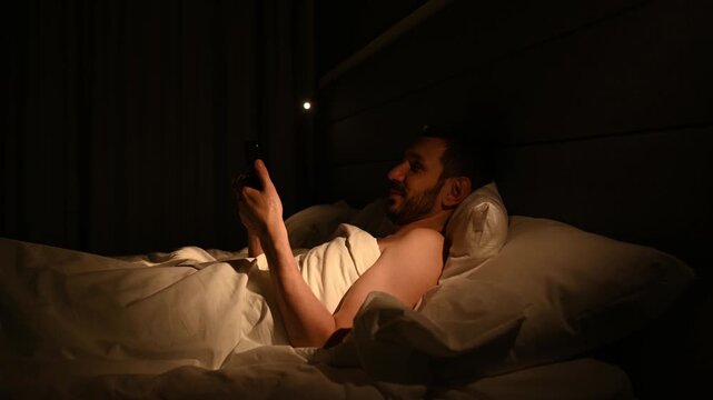 Smiling man lying in a dark bedroom at night, using his smartphone for social media, messaging, or watching videos, illuminated by the bright screen light before going to sleep