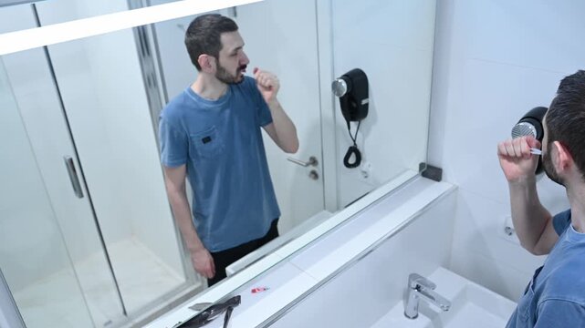 Millennial bearded man looking in the mirror while brushing his teeth with a toothbrush and toothpaste, taking care of his oral hygiene as part of his daily morning and evening routine