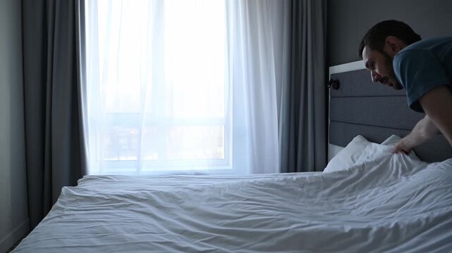 Young man diligently making his bed in a hotel room or apartment bedroom, arranging the white linens and fluffing the pillows as part of his daily morning routine and household chores