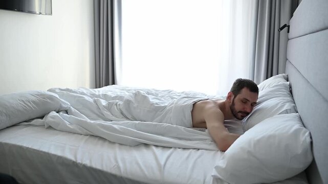 Young man sleeping peacefully in his comfortable bed in the morning, turning over in his sleep and changing position while dreaming. He is resting alone in a bright, modern bedroom