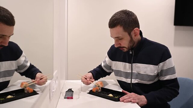 Hungry caucasian man with a beard sitting at a white table and eating delicious fresh sushi rolls from a takeaway container with chopsticks, enjoying his lunch break in a minimalist canteen