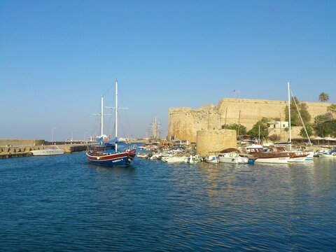 View of the historic Kyrenia Castle and the vibrant marina filled with various boats, including the wooden tourist gulet Sabrina II, under a clear blue sky. Kyrenia, Northern Cyprus. 2014. 