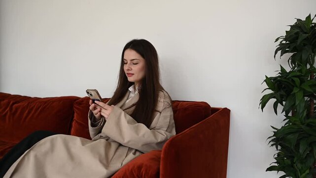 Young brunette woman in a beige trench coat sitting comfortably on a red velvet sofa in a modern living room, scrolling, texting, and browsing on her smartphone with a relaxed expression