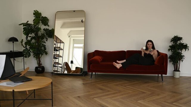 Young woman relaxing barefoot on a comfortable red sofa in a modern living room, drinking coffee and using her smartphone to scroll through social media on a peaceful morning