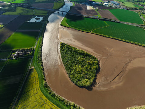Aerial view of Howden Dyke Island in the River Ouse surrounded by mudflats and lush green agricultural fields under bright sunlight in Goole, England, United Kingdom.