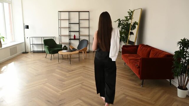 Graceful young woman with long brunette hair walking barefoot through her spacious, modern living room to enjoy a peaceful morning coffee, creating a serene and relaxing start to her day