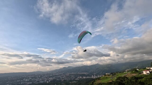 Paraglider flying over a city and mountain landscape under a blue sky with white clouds at sunset