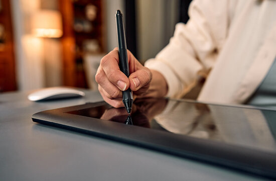 Close-up of hand holding stylus pen drawing on a graphics tablet in a cozy workspace.