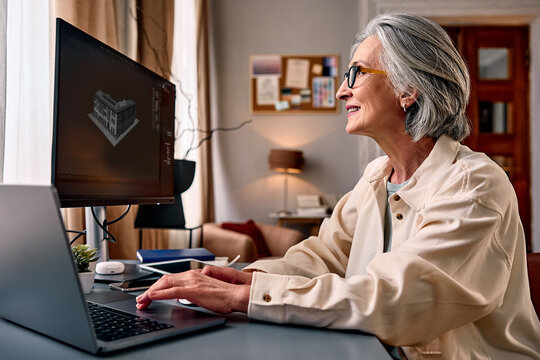 Senior woman working at home on laptop and external monitor in cozy living room, smiling while using computer for remote work, online learning, or digital design.