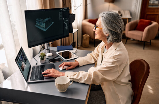 Senior woman working at a home office desk with dual monitors, laptop and tablet, designing a 3d architectural model on screen.