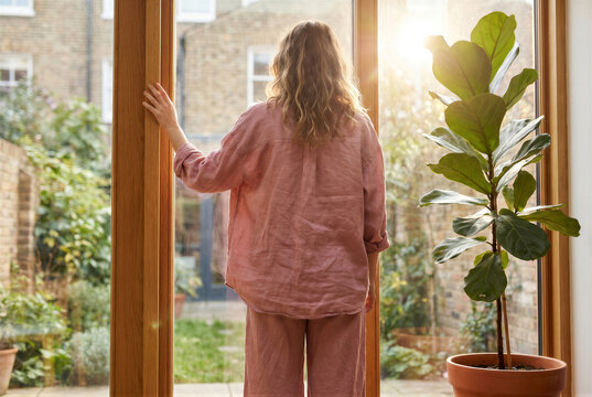 Woman standing by a large window looking out at a garden, wearing pink linen pajamas, back view, natural light, home interior