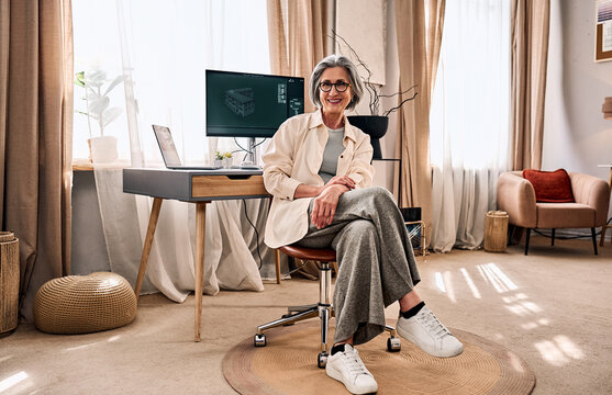A modern mature woman sits on a chair in a home office studio, looking at the camera and smiling. 