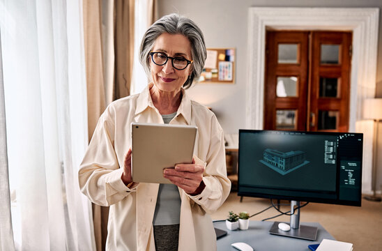 Senior female architect or engineer using tablet in home office with 3d building model on computer monitor, daylight interior, remote work concept.