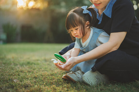 Curious Asian toddler girl and mother exploring nature with a magnifying glass in a garden at sunset. Cinematic golden hour shot of family bonding, educational play, and childhood discovery.