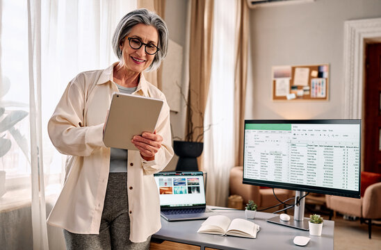 Smiling senior woman working from home, holding tablet beside desk with laptop and monitor showing spreadsheet, bright modern home office, remote work and productivity concept.