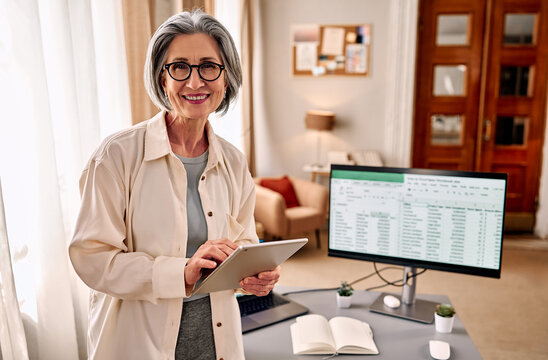 Confident middle-aged woman standing with a digital tablet in her home office.