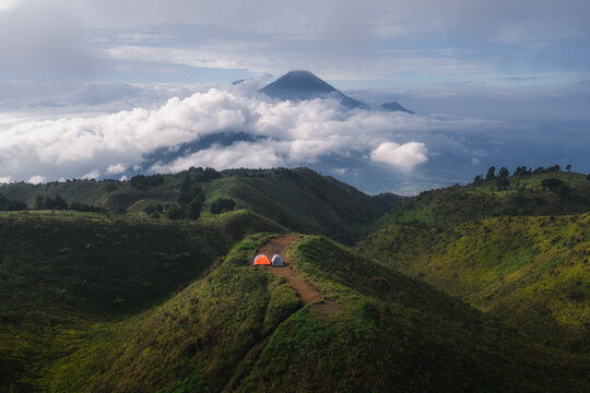 Aerial view of Gunung Prau ridge featuring camping tents on a trail overlooking rolling green hills and a distant volcanic peak above thick clouds in Mount Prau, Central Java, Indonesia.
