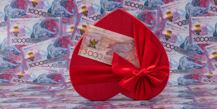 A 5,000 tenge banknote and a gift box in the shape of a human heart against a background of 10,000 tenge banknotes.
