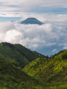 Aerial view of Gunung Prau peak rising above a thick sea of white clouds with lush green mountain ridges in the foreground under a soft sky Mount Prau, Central Java, Indonesia.