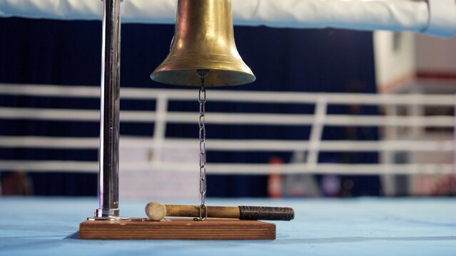 Golden boxing bell hanging from a chrome stand on a blue canvas, with a wooden mallet ready for signaling the start and end of rounds in a combat sport ring