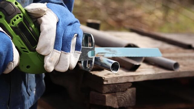Cordless reciprocating saw cuts metal pipe. A craftsman in protective gloves uses a cordless reciprocating saw (electric knife) to saw a thin pipe. Working with a power tool. Close-up of work