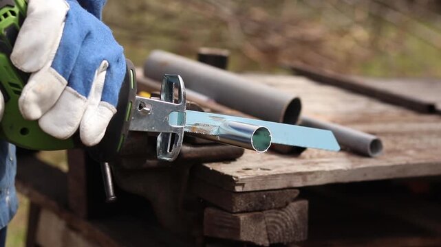 Cordless reciprocating saw cuts metal pipe. A craftsman in protective gloves uses a cordless reciprocating saw (electric knife) to saw a thin pipe. Working with a power tool. Close-up of work