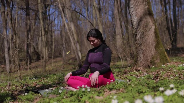 Young woman in comfortable sportswear practices yoga sitting in lotus pose on green lawn in forest. Concentrated female slowly tilts head forward and backward during calm exercise routine