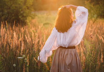 free-spirited woman in boho outfit walking away through golden meadow at sunset holding straw hat and enjoying serenity of nature during peaceful evening stroll in countryside landscape © Soloviova Liudmyla