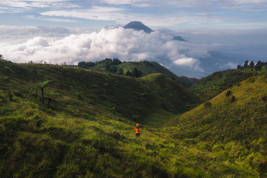 Aerial view of Gunung Prau with a hiker in an orange jacket overlooking lush green hills and a sea of clouds with a volcanic peak in the distance at Mount Prau, Central Java, Indonesia.