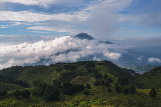 Aerial view of Gunung Prau with lush green hills and a thick sea of clouds under a blue sky in Mount Prau, Central Java, Indonesia.