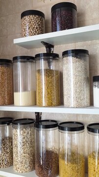 Pantry shelves displaying numerous transparent plastic containers filled with a variety of dry food ingredients, including grains, legumes, pulses, pasta, and cereals, for organized kitchen storage