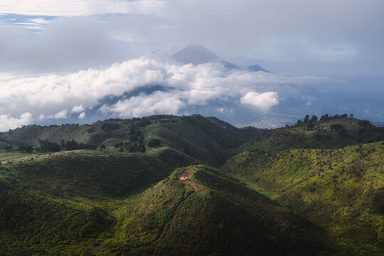 Aerial view of Gunung Prau with green rolling hills, small tents on a ridge, and a distant volcano peak rising above thick white clouds Mount Prau, Central Java, Indonesia.