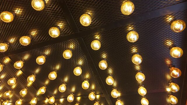 Rows of round vintage light bulbs flicker and blink on a black metal grid ceiling. Warm golden light creates a rhythmic patterns in a modern industrial loft style interior.