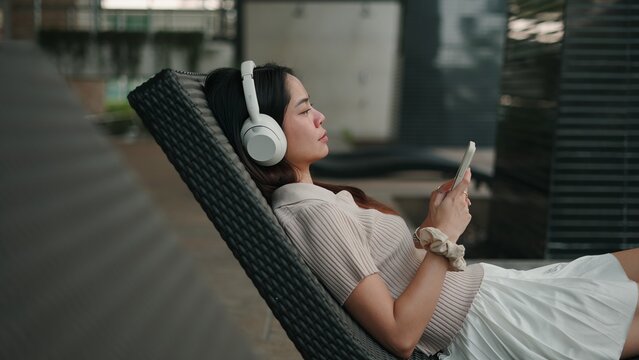 Young woman listening to music via wireless headphones, sitting on cozy lounge chair, scrolling smartphone screen outdoors