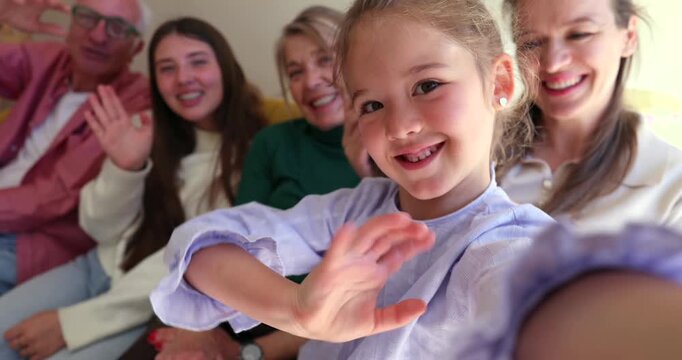 Little girl holding a camera during a video call with her family, including grandparents, mother, and aunt, all smiling and waving hello from the living room couch, capturing a joyful moment