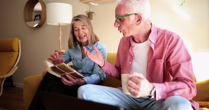 Elderly couple spending quality time together in their living room, the woman looking through a photo album while the man uses a laptop and drinks coffee, both smiling and talking