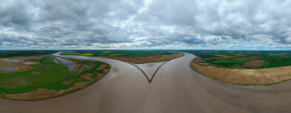 Aerial view of Trent Falls confluence where the River Trent and River Ouse meet, surrounded by green fields and yellow rapeseed under a cloudy sky in Goole, United Kingdom.