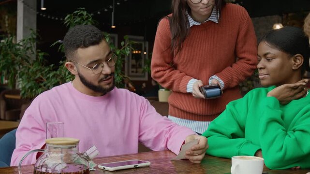 Gen Z man looking at restaurant bill and giving money to waitress after finishing lunch with diverse friends in cafe