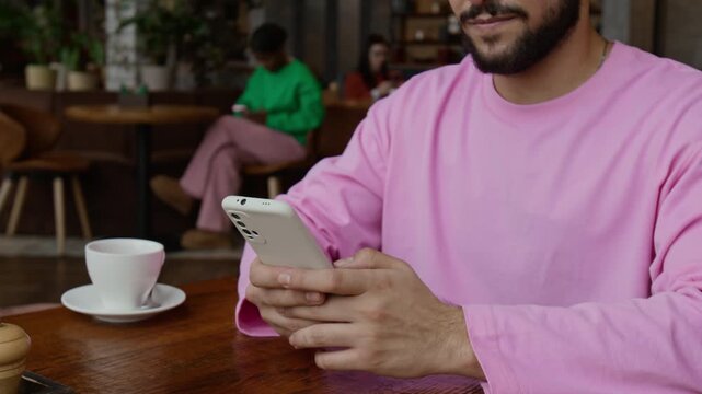 Midsection shot of bearded gen Z guy dressed in pink long-sleeved t-shirt texting on smartphone while sitting at cafe table with empty white cup on it