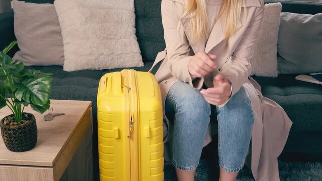 An anxious woman sit on sofa in living room with luggage, nervously check time on smartwatch while wait for taxi to go to airport.Concept of pre-travel anxiety, time pressure, departure preparation.
