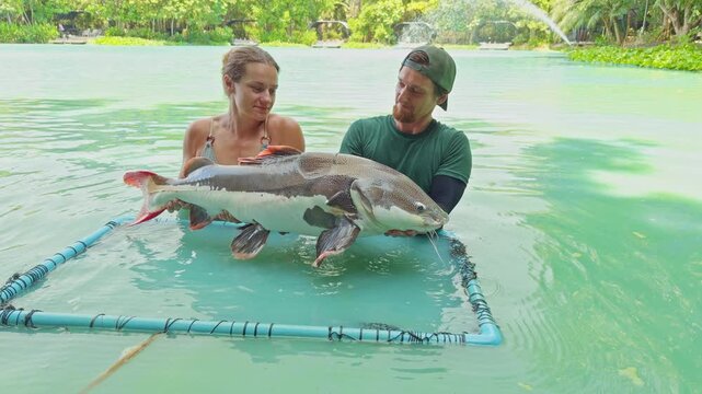 Couple Holding Large Redtail Catfish Together In Shallow Lake Water Inside Landing Frame With Smiles, Shared Fishing Experience And Leisure Activity