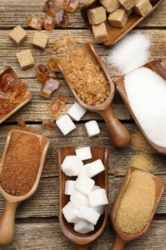 Different types of sugar in scoops on wooden table, flat lay