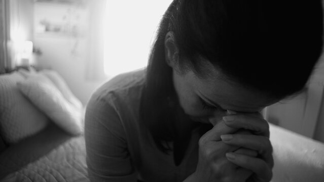 Close-up of woman sitting with head bowed and hands clasped, expressing emotional exhaustion, reflection, and quiet despair in intimate home setting, black and white